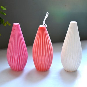 May include: Three teardrop-shaped candles in shades of pink, coral, and white. Each candle has a textured, geometric design and a wick. The candles are arranged on a neutral surface, creating a simple and elegant display.