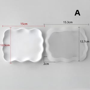 May include: Two white, wavy-edged ceramic plates. One measures 15 cm x 12.5 cm, the other 15.3 cm x 12.7 cm, with a 2 cm depth. The plates have a smooth, minimalist design, suitable for serving food or as decorative pieces.