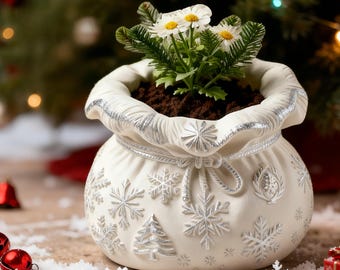 May include: A white ceramic Christmas-themed planter shaped like a bag, decorated with silver snowflakes and a tree. The planter holds soil and a small plant with white flowers and green leaves. The background features a blurred Christmas tree.