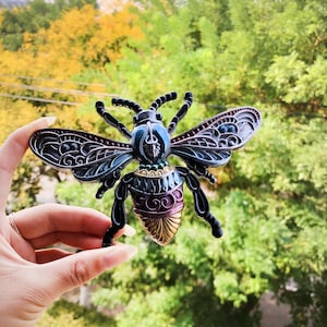 May include: A detailed, metal bee figurine with intricate wing and body designs. The bee features a combination of black, blue, gold, and purple colors. The wings have a filigree pattern, and the body has a layered design. The figurine is held against a blurred green and yellow background.