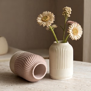 May include: Two ceramic vases, one upright and one on its side. The upright vase is off-white with vertical ridges and holds flowers. The other vase is pink with a wavy texture on the top half and off-white on the bottom.