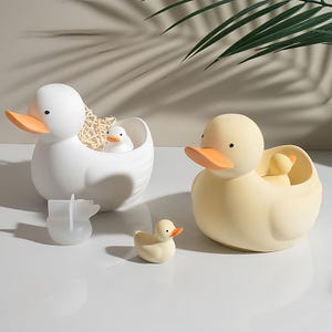 Op de afbeelding: Twee eendvormige containers, één wit en één geel, elk met een kleiner eendje. De witte eend heeft een eendje genesteld in stro. Er zijn ook een klein eendje en een doorzichtige, eendvormige mal aanwezig. Alle eenden hebben oranje snavels.