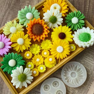 May include: A wooden tray displaying a collection of colourful, handmade flower-shaped soaps. The soaps are in shades of yellow, green, white, orange, and pink. Silicone moulds with flower designs are also present. The soaps are arranged to resemble a garden.