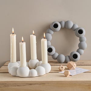 May include: A white candle holder with four lit candles and a gray ring-shaped candle holder. The white candle holder has a cloud-like design. Two small wooden geometric shapes and a piece of paper are also visible.