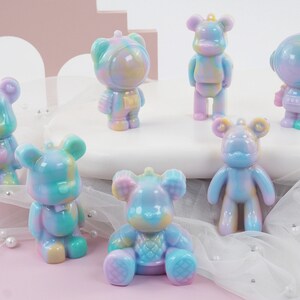K&ouml;nnte beinhalten: Eine Sammlung farbenfroher B&auml;renfiguren in verschiedenen Posen. Die B&auml;ren sind haupts&auml;chlich Pastellt&ouml;ne von Blau, Lila, Gelb und Gr&uuml;n. Einige stehen, w&auml;hrend andere sitzen oder Astronautenhelme tragen. Die Figuren haben ein gl&auml;nzendes Finish.