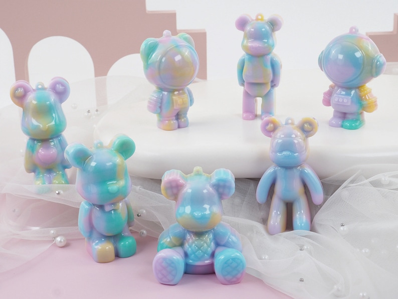 K&ouml;nnte beinhalten: Eine Sammlung farbenfroher B&auml;renfiguren in verschiedenen Posen. Die B&auml;ren sind haupts&auml;chlich Pastellt&ouml;ne von Blau, Lila, Gelb und Gr&uuml;n. Einige stehen, w&auml;hrend andere sitzen oder Astronautenhelme tragen. Die Figuren haben ein gl&auml;nzendes Finish.