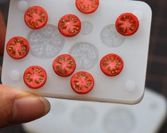 May include: A close-up of a white silicone mold filled with miniature, red tomato slices. The mold has several circular indentations, and the tomato slices are arranged on top. The tomatoes have a detailed, realistic appearance.