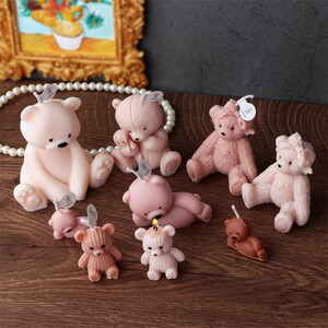 May include: A collection of teddy bear-shaped candles in various shades of pink and brown. Some bears are sitting, lying down, or have textured fur. Each candle has a small tag attached. The candles are arranged on a wooden surface.