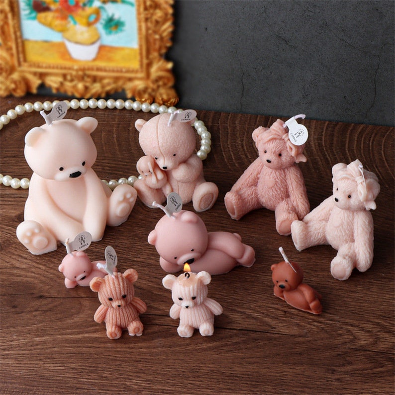 May include: A collection of teddy bear-shaped candles in various shades of pink and brown. Some bears are sitting, lying down, or have textured fur. Each candle has a small tag attached. The candles are arranged on a wooden surface.