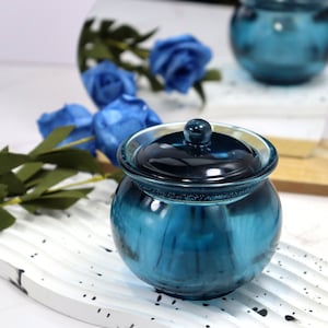 Op de afbeelding: Een teal glazen pot met een deksel. De pot is rond en heeft een glanzende afwerking. Het deksel heeft een kleine knop. De pot staat op een wit, golvend oppervlak. Blauwe rozen zijn op de achtergrond.