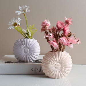 Op de afbeelding: Twee decoratieve vazen, één licht lavendelkleurig en één lichtroze, elk met bloemen. De vazen hebben een textuur met een zonnestraalontwerp. De lavendelvaas bevat witte madeliefjes en de roze vaas bevat roze gedroogde bloemen.