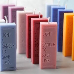 Puede incluir: Una colección de velas rectangulares en varios colores, incluyendo blanco, lavanda, rojo, rosa, azul y naranja. Cada vela tiene una mecha blanca y el texto "LIGHT A CANDLE TO SAVE TIME" impreso.