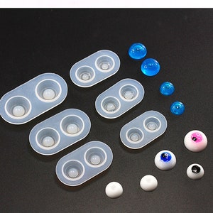 May include: A collection of silicone molds for crafting doll eyes, along with various finished eye pieces. The molds are translucent, and the finished eyes are white, blue, and pink. Some eyes have black pupils.
