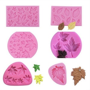 May include: A collection of pink silicone molds in various shapes, featuring leaf designs. The molds include rectangular, oval, and triangular shapes, with detailed leaf impressions. Several finished leaf-shaped decorations in brown, green, and yellow are also shown.