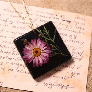 May include: A square pendant necklace with a gold chain. The black pendant features a pressed pink daisy with a yellow centre, green leaves, and buds. It rests on a vintage paper background with handwritten text.