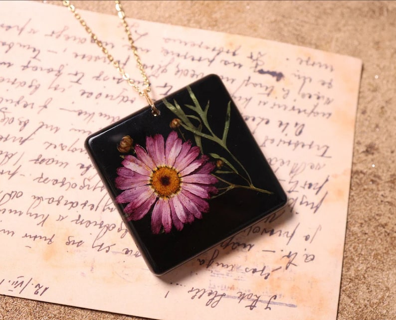 May include: A square pendant necklace with a gold chain. The black pendant features a pressed pink daisy with a yellow centre, green leaves, and buds. It rests on a vintage paper background with handwritten text.