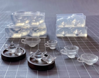 May include: Clear resin miniature tea sets, including teapots, cups, and saucers, are displayed on a gray cutting mat. Two sets are on dark brown trays. Several molds for the tea sets are also visible.