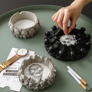 Puede incluir: Tres ceniceros decorativos con diseños de calaveras. Uno es negro, otro gris y el tercero también gris. Los ceniceros están llenos de una sustancia blanca. Una mano coloca un cigarrillo en el cenicero negro.