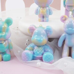 K&ouml;nnte beinhalten: Eine Sammlung bunter B&auml;renfiguren in Pastellt&ouml;nen von Blau, Gelb und Lila. Einige stehen, andere sitzen, und einer ist als Astronaut verkleidet. Jede Figur hat eine Schlaufe zum Aufh&auml;ngen.