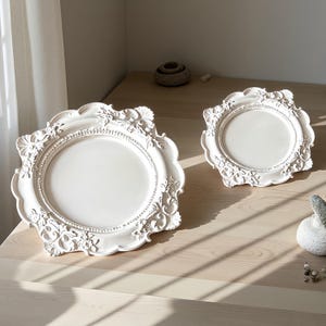 Puede incluir: Dos platos decorativos circulares de color blanco roto con intrincados relieves de flores y volutas a lo largo de los bordes festoneados. Los platos tienen un centro liso y mate y un borde texturizado y ornamentado con un diseño clásico.