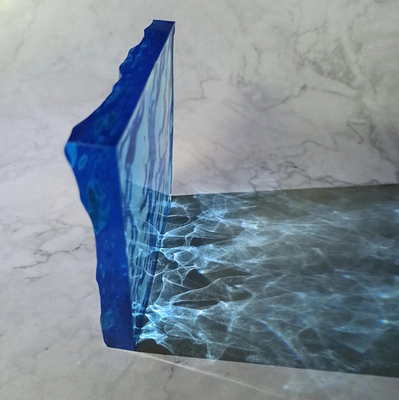 May include: A blue resin bookend with a textured, irregular edge. The bookend casts a shadow with a rippling water effect. The color of the bookend is a deep blue with lighter blue marbling.