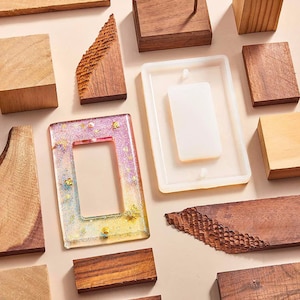 Puede incluir: Vista aérea de suministros para trabajos en madera y resina. Un molde rectangular de resina con un inserto blanco se encuentra junto a un marco de resina colorido con purpurina y pan de oro. Bloques de madera de diferentes tamaños y texturas están dispuestos.