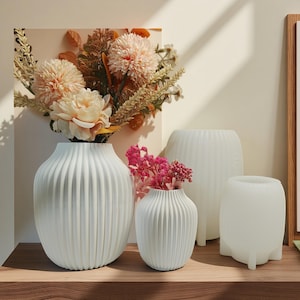 Pode incluir: Uma coleção de quatro vasos de cerâmica branca com detalhes verticais canelados. Os vasos variam em tamanho e forma, alguns contêm flores secas em tons de rosa, pêssego e creme. Os vasos são exibidos em uma superfície de madeira.