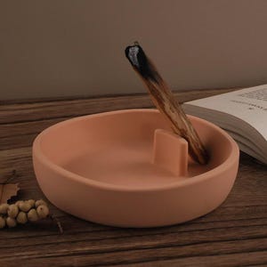 Può includere: Un porta incenso in ceramica color terracotta con un supporto rettangolare. Un bastoncino di Palo Santo acceso è appoggiato nel supporto. Il supporto è rotondo con una forma a ciotola poco profonda. Un libro è aperto sullo sfondo.