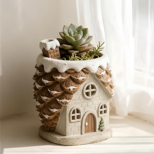 May include: A house-shaped plant pot with a succulent plant. The pot features a white brick facade, a brown wooden door, and a chimney. The roof is covered in brown shingles with white accents, and the top is trimmed with white. A small green Christmas tree is next to the door.