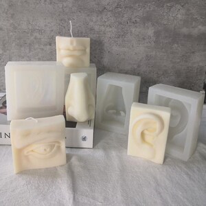 May include: A collection of ivory-colored candles and molds, each shaped like human facial features: lips, a nose, an eye, and an ear. The candles are square, with a wick on top. The molds are translucent and show the negative space of the features.