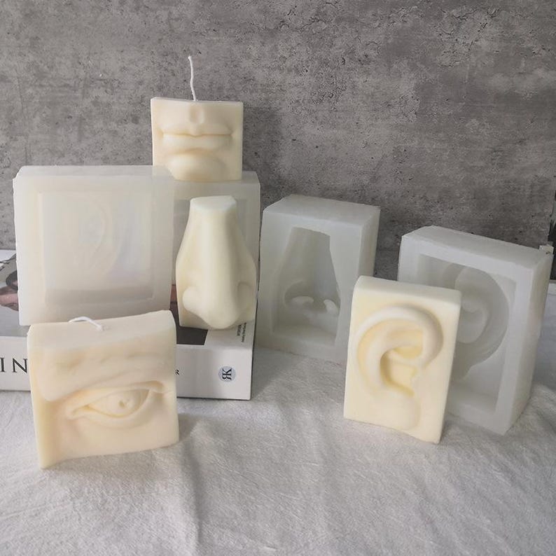 May include: A collection of ivory-colored candles and molds, each shaped like human facial features: lips, a nose, an eye, and an ear. The candles are square, with a wick on top. The molds are translucent and show the negative space of the features.