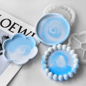 Può includere: Una collezione di vassoi in resina di varie forme e design. I vassoi presentano un motivo a spirale blu e bianco. Un vassoio è a forma di nuvola, un altro è rotondo e un terzo ha un bordo perlato. La parola "LOEWE" è parzialmente visibile.