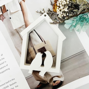 Könnte beinhalten: Ein weißer, hausförmiger Rahmen mit zwei weißen Vogelsilhouetten. Im Inneren des Rahmens befindet sich eine gewebte Tragetasche mit braunen Ledergriffen und dem Markennamen "LOEWE". Der Hintergrund umfasst getrocknete Blumen und eine Person in beiger Kleidung.