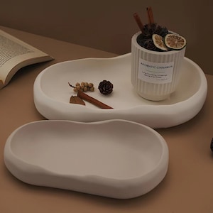 Könnte beinhalten: Zwei weiße, organisch geformte Tabletts. Das obere Tablett enthält einen weißen, gerippten Keramikbehälter mit der Aufschrift "AROMATIC CINNAMON" und getrockneten Pflanzen. Zimtstangen, Tannenzapfen und getrocknete Zitrusfrüchte sind auf den Tabletts angeordnet.