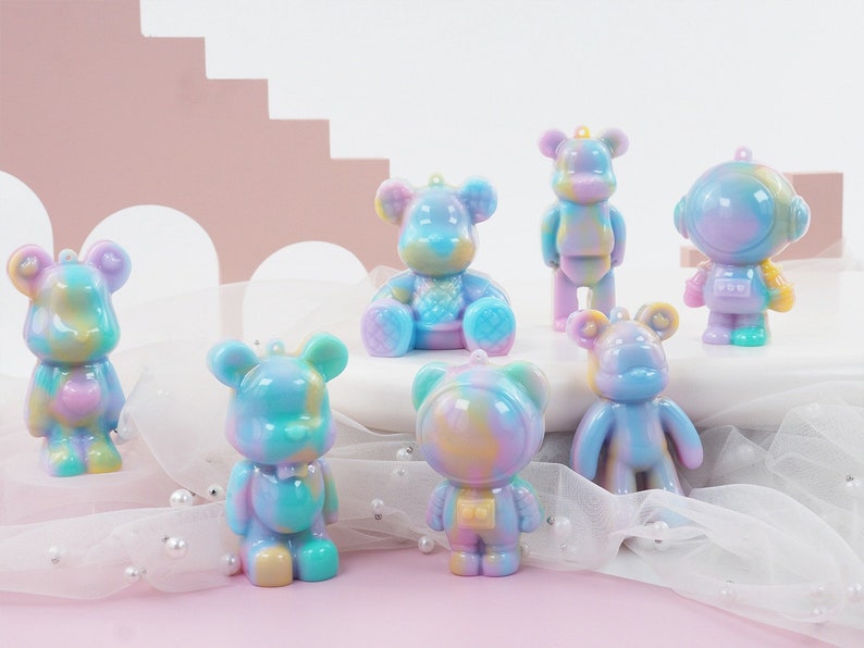 K&ouml;nnte beinhalten: Eine Sammlung farbenfroher B&auml;renfiguren in verschiedenen Posen. Die Figuren weisen einen Batik-Effekt mit Pastellt&ouml;nen von Blau, Lila, Gelb und Gr&uuml;n auf. Eine Figur ist als Astronaut gestaltet. Jede hat eine kleine Schlaufe zum Aufh&auml;ngen.