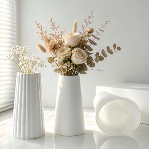 Puede incluir: Dos floreros de cerámica blanca y uno acostado de lado. Un florero tiene una textura acanalada vertical, mientras que el otro es liso y cónico. Los floreros verticales contienen arreglos de rosas beige secas, eucalipto y flores blancas.