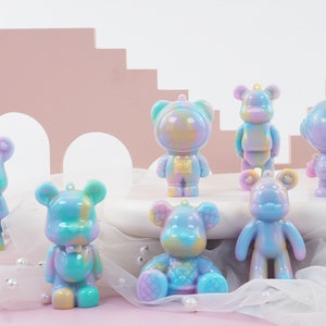 K&ouml;nnte beinhalten: Eine Sammlung farbenfroher B&auml;renfiguren in verschiedenen Posen. Die B&auml;ren sind haupts&auml;chlich in Pastellt&ouml;nen von Blau, Lila, Gelb und Gr&uuml;n gehalten. Einige B&auml;ren tragen Astronautenanz&uuml;ge. Jeder B&auml;r hat oben eine kleine Schlaufe zum Aufh&auml;ngen.
