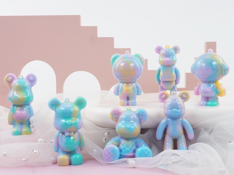 K&ouml;nnte beinhalten: Eine Sammlung farbenfroher B&auml;renfiguren in verschiedenen Posen. Die B&auml;ren sind haupts&auml;chlich in Pastellt&ouml;nen von Blau, Lila, Gelb und Gr&uuml;n gehalten. Einige B&auml;ren tragen Astronautenanz&uuml;ge. Jeder B&auml;r hat oben eine kleine Schlaufe zum Aufh&auml;ngen.