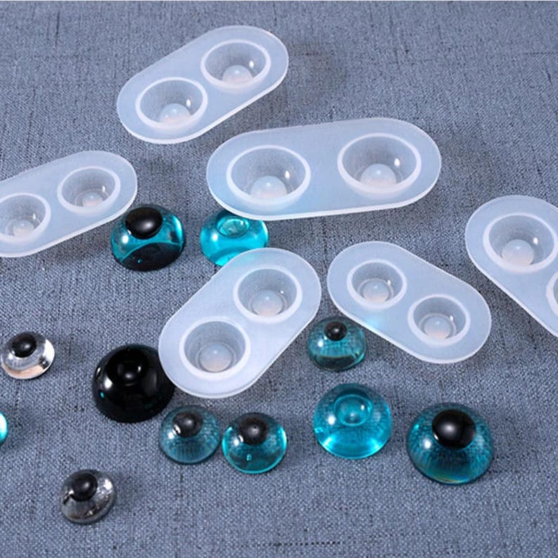 May include: A collection of clear silicone molds with two circular cavities each, alongside various glass eye cabochons in shades of blue, black, and silver. The cabochons have a domed shape and a reflective quality, suggesting they are for crafting.