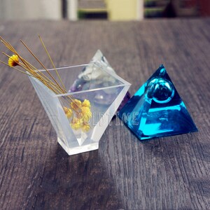 May include: Clear, geometric resin pyramid-shaped containers. One container holds dried yellow flowers and thin stems. A blue pyramid with a bubble is to the right. The word "MOLDTIME" is visible on the clear container.