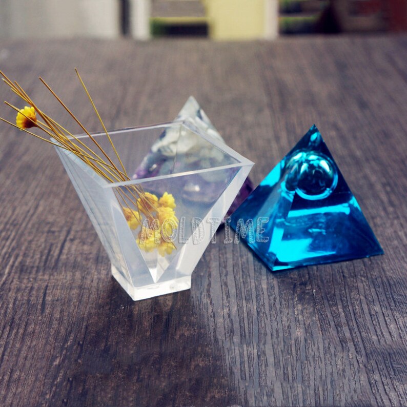 May include: Clear, geometric resin pyramid-shaped containers. One container holds dried yellow flowers and thin stems. A blue pyramid with a bubble is to the right. The word "MOLDTIME" is visible on the clear container.