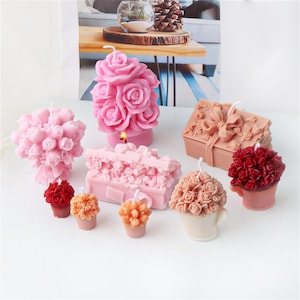 May include: A selection of decorative candles in various shapes and colours. The candles include a large pink rose-shaped candle, a bouquet of pink roses, a gift box, and smaller candles in the shape of flowers. The candles are on a white surface.