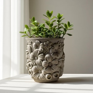 May include: A gray, floral-patterned planter pot filled with a green leafy plant. The pot has a detailed, embossed design of various flowers. The plant's green leaves contrast with the pot's neutral color, creating a visually appealing display.