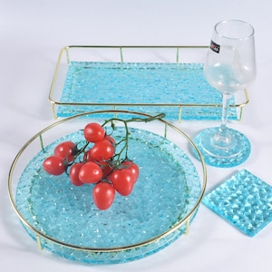 May include: A selection of serving items, including a round tray, rectangular tray, coaster, and a wine glass. The trays and coaster are a textured, light blue glass with gold-coloured metal rims. A cluster of red tomatoes sits on the round tray.