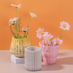 May include: Three decorative flower pots in yellow, pink, and gray, each holding artificial flowers. The yellow pot has a textured design, the pink pot has a geometric pattern, and the gray pot has vertical lines. The background is orange.