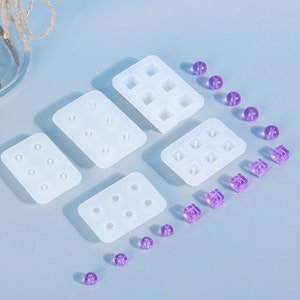 May include: A collection of white silicone molds in various rectangular shapes, designed for crafting. The molds are accompanied by small, translucent purple beads and cube-shaped embellishments, ideal for jewelry making or other DIY projects.