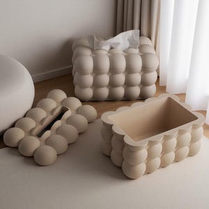 Puede incluir: Conjunto de tres contenedores de almacenamiento decorativos beige con diseño de burbujas. El conjunto incluye una caja de pañuelos, una bandeja y una caja rectangular. Los artículos son de material liso y sólido, colocados sobre una alfombra clara y suelo de madera.