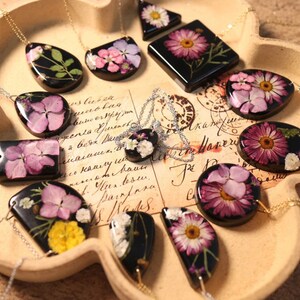 May include: A selection of resin necklaces with pressed flowers. Each pendant, in various shapes, displays a different floral arrangement on a black base, suspended from a delicate silver chain. The necklaces are displayed on a tray.
