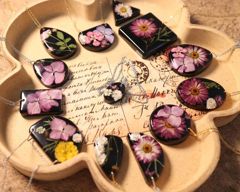 May include: A selection of resin necklaces with pressed flowers. Each pendant, in various shapes, displays a different floral arrangement on a black base, suspended from a delicate silver chain. The necklaces are displayed on a tray.