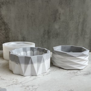 Może przedstawiać: Trzy geometryczne donice betonowe w odcieniach szarości i bieli. Donice mają fasetowane wzory i różne wysokości. Jedna to forma. Tło jest nakrapiane szarością.